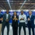 Five professionals in business attire give thumbs up at a car showroom/event, smiling in front of a white vehicle and blue banners.