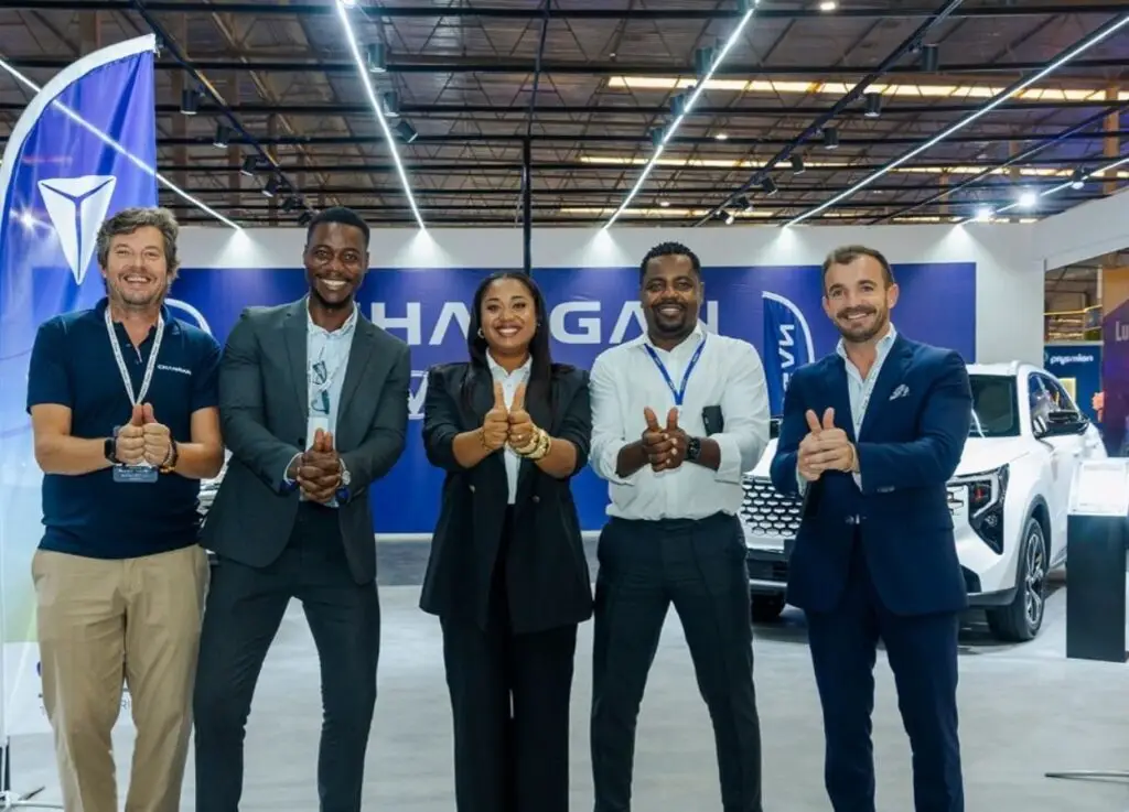 Five professionals in business attire give thumbs up at a car showroom/event, smiling in front of a white vehicle and blue banners.