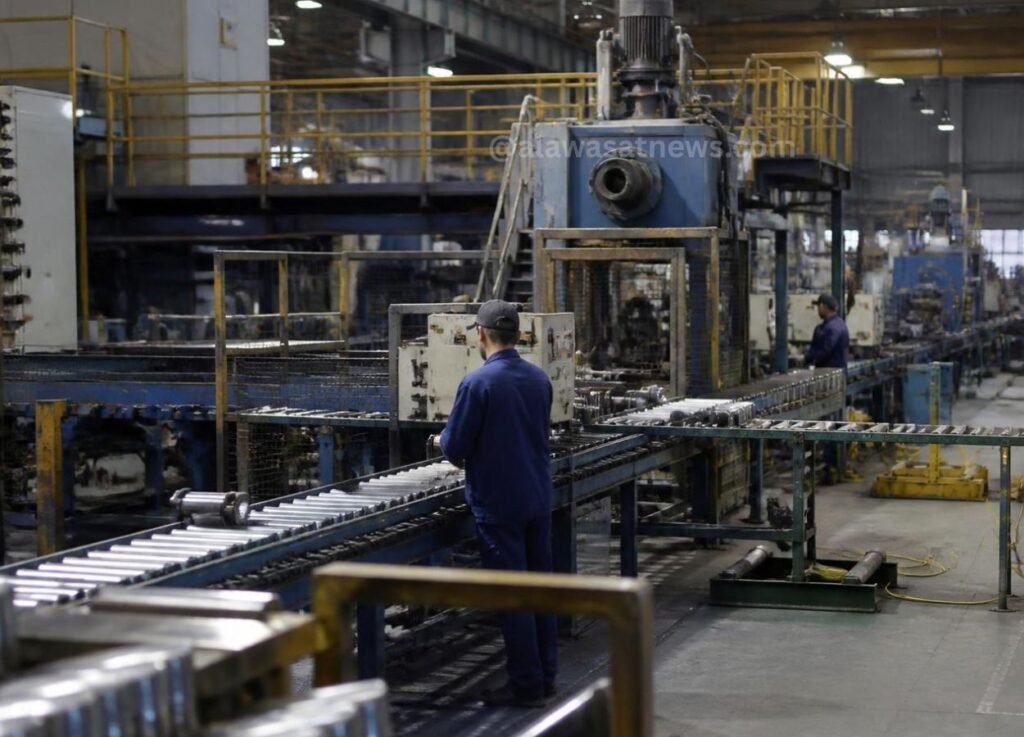 Industrial factory floor with a roller conveyor and large blue press; worker in blue coveralls stands at a control panel.