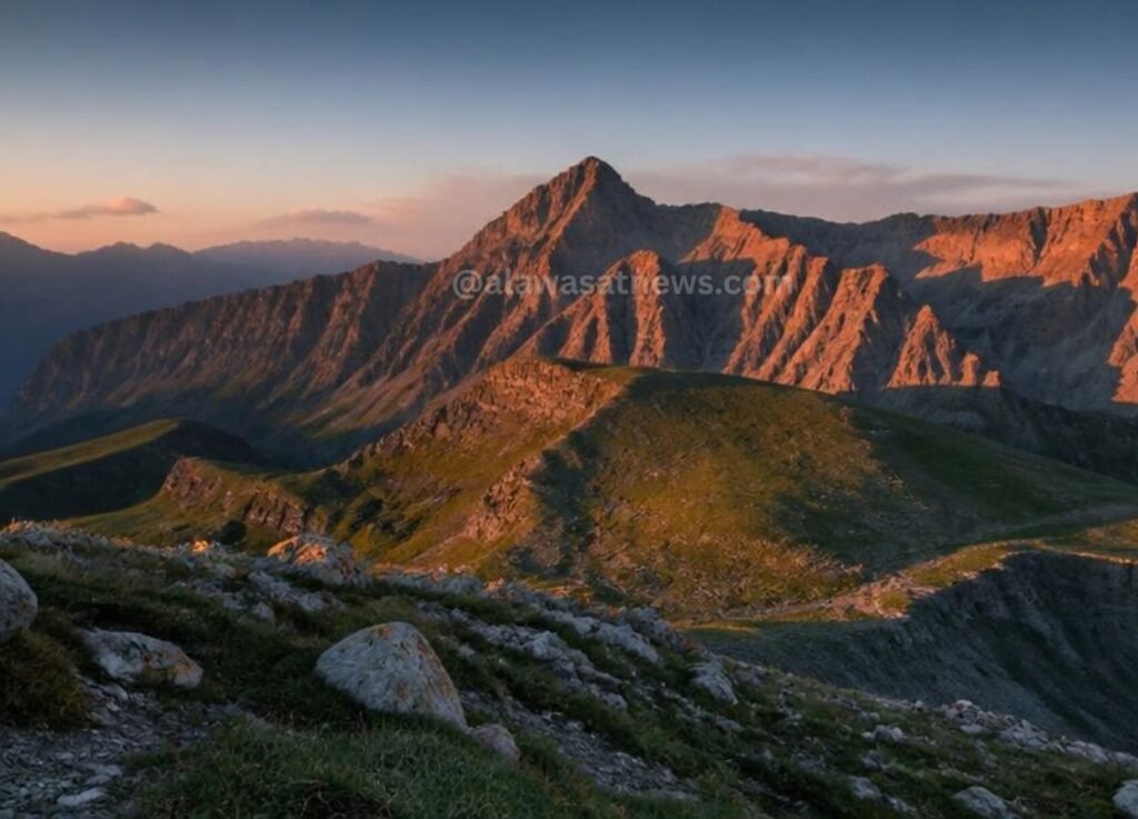 Sunlit jagged mountain peaks at sunset with a rocky foreground and green slopes.