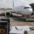 Airplane at a jet bridge being serviced on the tarmac with fuel barrels, paperwork, a calculator, and coins in the foreground used for illustrating aviation operations and refueling planning.